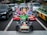 A group of people dressed in colorful costumes, inspired by popular video game characters, are riding go-karts on a busy city street. The go-karts are brightly decorated and situated among regular cars, waiting at a traffic light.