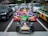 A group of people dressed in colorful costumes, inspired by popular video game characters, are riding go-karts on a busy city street. The go-karts are brightly decorated and situated among regular cars, waiting at a traffic light.