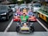 A group of people dressed in colorful costumes, inspired by popular video game characters, are riding go-karts on a busy city street. The go-karts are brightly decorated and situated among regular cars, waiting at a traffic light.