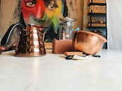 A tabletop scene featuring a variety of kitchenware, including a copper kettle, a metal bowl, and a glass jar. In the background, a vibrant, colorful mural of a face adds an artistic element. To the right are shelves lined with small boxes and a brick placed among the objects.