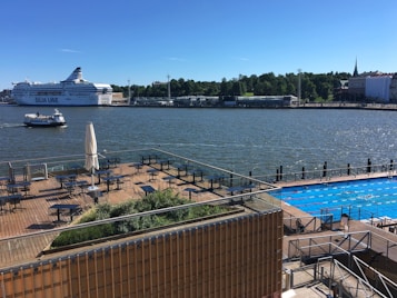 A large cruise ship labeled 'Silja Line' is sailing on a calm body of water, possibly a bay or river. There is a smaller boat nearby. In the foreground, there's a wooden deck seating area with tables, chairs, and a closed umbrella. An outdoor swimming pool with lanes is visible on the right. The background shows a densely forested area and some buildings, with a clear blue sky.