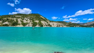 A serene body of turquoise water stretches out under a bright blue sky dotted with fluffy white clouds. In the distance, a person is stand-up paddleboarding near the shoreline, surrounded by lush green foliage and rocky hillsides. The peaceful scene captures a sense of tranquility and natural beauty.