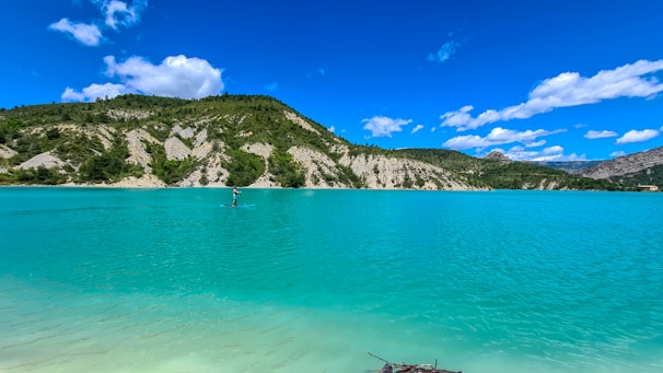 A serene body of turquoise water stretches out under a bright blue sky dotted with fluffy white clouds. In the distance, a person is stand-up paddleboarding near the shoreline, surrounded by lush green foliage and rocky hillsides. The peaceful scene captures a sense of tranquility and natural beauty.