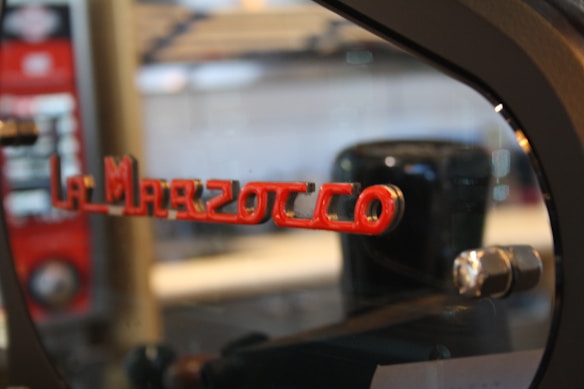 A close-up view of a metal espresso machine with the name La Marzocco in red, cut-out letters. The machine has a reflective surface, with visible mechanical components. Blurred background elements suggest an indoor setting.