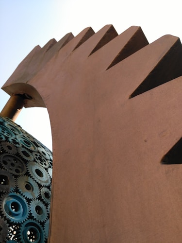 A large, abstract metal sculpture featuring a curved structure with jagged edges and a base composed of numerous interconnected metallic gears. The sculpture is set against a clear, pale blue sky, highlighting the texture and industrial design.