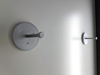 Two metallic wall hooks are mounted on a plain, light-colored wall. Each hook has a circular base with visible screws and a protruding cylindrical peg.