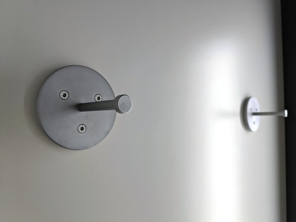 Two metallic wall hooks are mounted on a plain, light-colored wall. Each hook has a circular base with visible screws and a protruding cylindrical peg.