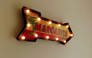 A retro-style arrow-shaped sign with light bulbs along the edges pointing to the right. The sign features a green margarita glass graphic and the words 'Margarita Bar' in bold, colorful letters.