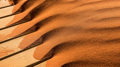 Waves of fine, reddish-brown sand create ripples and patterns across the surface of a wooden deck. The sand appears to have been blown naturally, forming parallel lines and gentle curves, accentuating textures against the light brown wood.