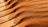 Waves of fine, reddish-brown sand create ripples and patterns across the surface of a wooden deck. The sand appears to have been blown naturally, forming parallel lines and gentle curves, accentuating textures against the light brown wood.