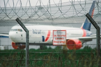 A large commercial airplane is positioned in the background behind a tall wire security fence topped with barbed wire. A red and white warning sign in a Cyrillic script is attached to the fence, signaling a restricted area. The environment appears to be an airport or aviation facility.