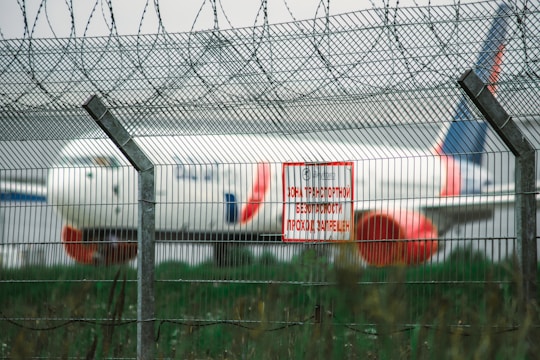 A large commercial airplane is positioned in the background behind a tall wire security fence topped with barbed wire. A red and white warning sign in a Cyrillic script is attached to the fence, signaling a restricted area. The environment appears to be an airport or aviation facility.