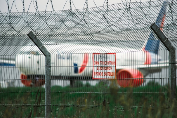 A large commercial airplane is positioned in the background behind a tall wire security fence topped with barbed wire. A red and white warning sign in a Cyrillic script is attached to the fence, signaling a restricted area. The environment appears to be an airport or aviation facility.