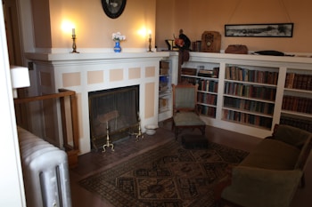 A cozy, vintage-style living room featuring a fireplace with lit candles on the mantel, a small blue vase with flowers, and bookshelves filled with a variety of books. A vintage radio and a framed photograph are also present on the shelf. A plush armchair and a small sofa sit on an intricately patterned rug, while a brass fireguard stands in front of the fireplace.