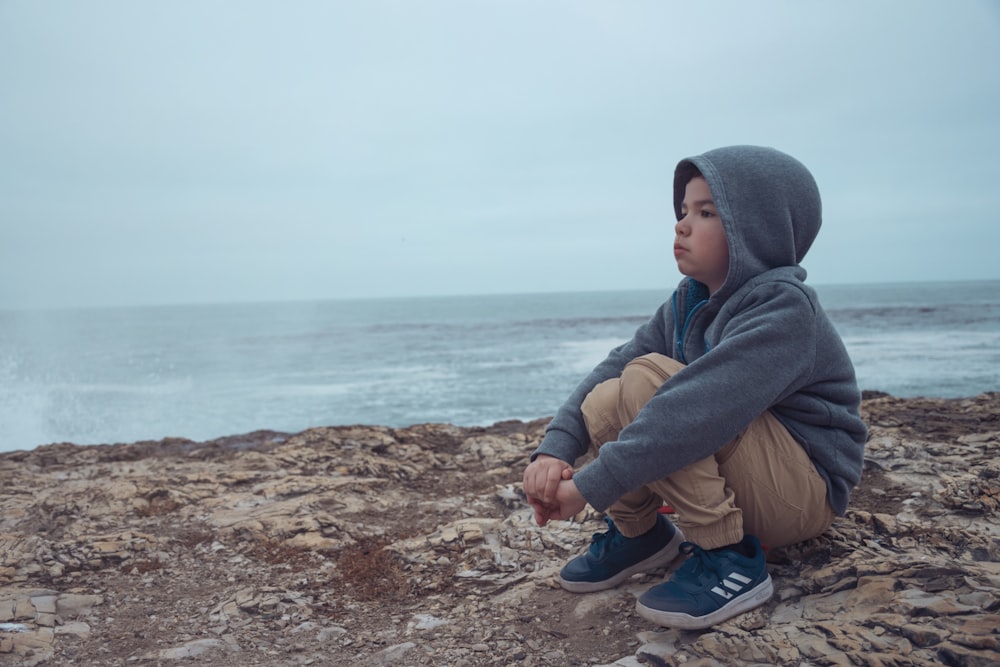 man in gray hoodie and brown pants sitting on brown rock near body of water during