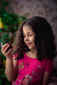 A young girl with long, dark hair wearing a bright pink shirt decorated with butterflies and heart motifs is holding a small fruit. She appears to be observing it with an expression of curiosity and delight. The background is soft-focused, featuring some green plants.