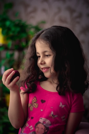 A young girl with long, dark hair wearing a bright pink shirt decorated with butterflies and heart motifs is holding a small fruit. She appears to be observing it with an expression of curiosity and delight. The background is soft-focused, featuring some green plants.