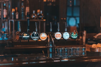 A row of beer taps is installed on a bar counter, showcasing different brands under dim, warm lighting. The setup includes glassware neatly arranged beneath the taps, and the background features blurred shelves with bottles, creating an intimate and relaxed atmosphere.