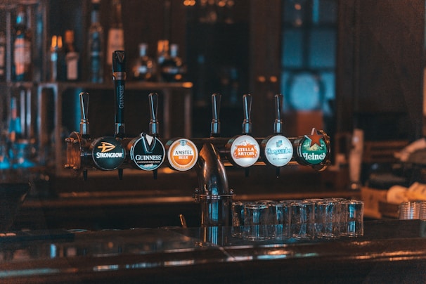 A row of beer taps is installed on a bar counter, showcasing different brands under dim, warm lighting. The setup includes glassware neatly arranged beneath the taps, and the background features blurred shelves with bottles, creating an intimate and relaxed atmosphere.