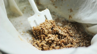 A close-up view of a white spatula stirring a mixture of wet grains and liquid, suggesting a process of brewing or cooking. The grains appear soaked and are contained within a light fabric, possibly cheesecloth, for straining purposes.