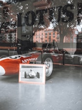 A classic red racing car, likely vintage, is displayed behind a glass window with the word 'LOTUS' visible on the window. A framed black and white photograph of a similar car is positioned in front of the vehicle. Reflections of outdoors, including trees and buildings, can be seen on the glass.