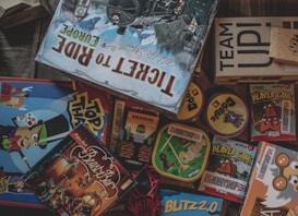 A collection of board game boxes with various designs and titles, including Ticket to Ride Europe, Top That!, and Beaver Gang. The games are stacked and arranged closely together, with colorful illustrations and text visible on the boxes. The setting suggests a playful and engaging environment, with hints of strategy and fun.