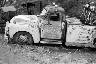 The black and white photograph features an old, rusted tow truck with a partially visible signage that reads 'Cannon's Salvage & Towing.' The truck is parked amidst a rugged, overgrown area filled with grass and gravel. The vehicle's condition suggests abandonment, with significant wear and tear visible. In the background, there are other old, dilapidated vehicles or machinery parts, adding to the vintage and neglected feel of the scene.