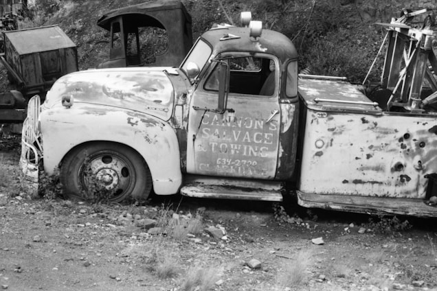 The black and white photograph features an old, rusted tow truck with a partially visible signage that reads 'Cannon's Salvage & Towing.' The truck is parked amidst a rugged, overgrown area filled with grass and gravel. The vehicle's condition suggests abandonment, with significant wear and tear visible. In the background, there are other old, dilapidated vehicles or machinery parts, adding to the vintage and neglected feel of the scene.
