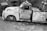 The black and white photograph features an old, rusted tow truck with a partially visible signage that reads 'Cannon's Salvage & Towing.' The truck is parked amidst a rugged, overgrown area filled with grass and gravel. The vehicle's condition suggests abandonment, with significant wear and tear visible. In the background, there are other old, dilapidated vehicles or machinery parts, adding to the vintage and neglected feel of the scene.