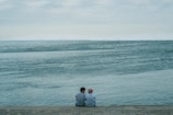 Two people sit together on a stone embankment, facing a vast, serene body of water under an overcast sky. Their closeness and the tranquil setting suggest a moment of quiet companionship.