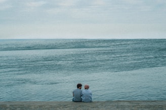 Two people sit together on a stone embankment, facing a vast, serene body of water under an overcast sky. Their closeness and the tranquil setting suggest a moment of quiet companionship.