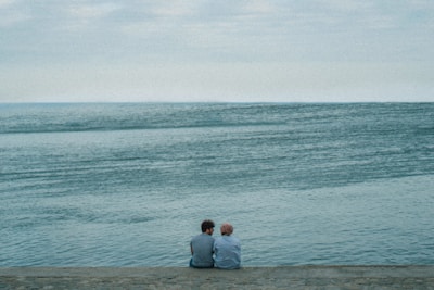 Two people sit together on a stone embankment, facing a vast, serene body of water under an overcast sky. Their closeness and the tranquil setting suggest a moment of quiet companionship.