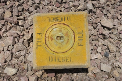 A square metal plate, painted yellow, is surrounded by gravel. The plate has a circular panel in the middle with the word 'Diesel' and 'Fill' written on it. The surface shows signs of weathering and rust.