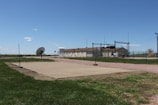 A fenced-in area with a large satellite dish and a few buildings in the background, set in a grassy landscape under a clear blue sky. There are poles around a rectangular sandy area and other structures, including a water tank and antennas.