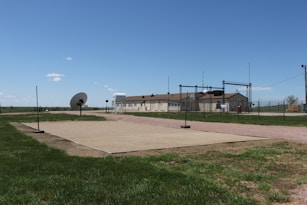 A fenced-in area with a large satellite dish and a few buildings in the background, set in a grassy landscape under a clear blue sky. There are poles around a rectangular sandy area and other structures, including a water tank and antennas.