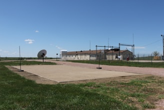 A fenced-in area with a large satellite dish and a few buildings in the background, set in a grassy landscape under a clear blue sky. There are poles around a rectangular sandy area and other structures, including a water tank and antennas.
