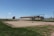 A fenced-in area with a large satellite dish and a few buildings in the background, set in a grassy landscape under a clear blue sky. There are poles around a rectangular sandy area and other structures, including a water tank and antennas.