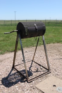 A cylindrical metal drum with a mesh surface mounted on a triangular stand with a hand crank on the side. It is positioned outdoors on a gravel area within a grassy landscape, fenced in the background under a clear blue sky.