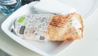 A wrapped sandwich on a white plastic plate with grill marks on the bread, partially unwrapped to show fillings of lettuce, sauce, and tomatoes. A water bottle and a napkin are visible in the background.