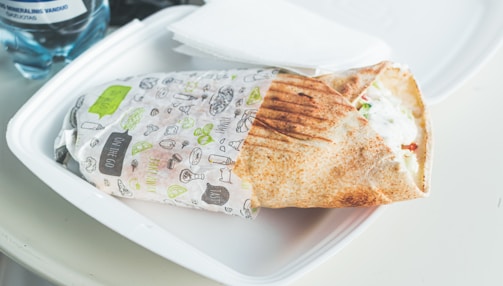A wrapped sandwich on a white plastic plate with grill marks on the bread, partially unwrapped to show fillings of lettuce, sauce, and tomatoes. A water bottle and a napkin are visible in the background.