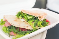 A sandwich made with pita bread filled with fresh green lettuce, slices of pink ham, and possibly some cheese. It is placed on a white disposable plate, likely a to-go container, against a blurred background.