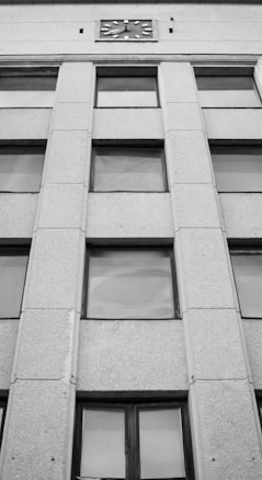 The image features the facade of a multi-story building with a symmetrical design. Vertical columns separate the rows of rectangular windows. At the top center, there is a clock embedded in the wall. The building's exterior is constructed from a textured material, giving it a robust appearance.