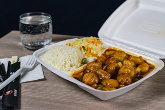 A takeout container filled with a meal consisting of rice, vegetables, and what appears to be chicken in sauce is placed on a light wooden table. Next to it, there is a glass of water. Also on the table are chopsticks, a plastic fork, and some napkins.