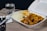A takeout container filled with a meal consisting of rice, vegetables, and what appears to be chicken in sauce is placed on a light wooden table. Next to it, there is a glass of water. Also on the table are chopsticks, a plastic fork, and some napkins.