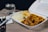 A takeout container filled with a meal consisting of rice, vegetables, and what appears to be chicken in sauce is placed on a light wooden table. Next to it, there is a glass of water. Also on the table are chopsticks, a plastic fork, and some napkins.