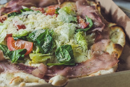 A freshly baked pizza topped with generous amounts of prosciutto, fresh spinach leaves, sliced tomatoes, and grated cheese. The crust is golden brown and crispy, forming a contrast with the colorful, vibrant toppings that are neatly arranged.