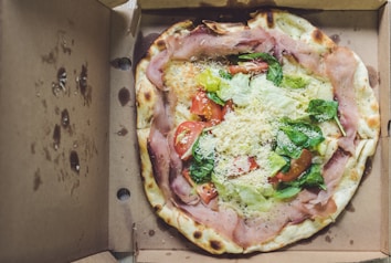 A pizza topped with ingredients like ham, fresh lettuce, spinach, tomato slices, and grated cheese sits inside a brown cardboard box. The crust is slightly charred, indicating it has been baked well. The box has some grease stains on the inside.