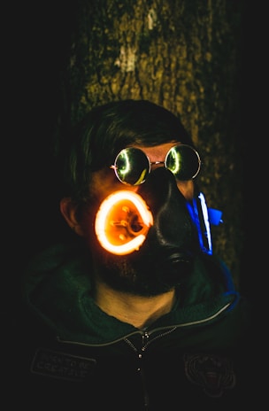 A person wearing round reflective sunglasses and a futuristic gas mask with glowing neon rings. The background is dark, making the glowing elements stand out. The person is dressed in a dark hoodie and jacket.