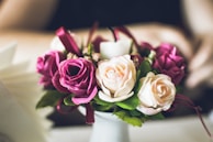 A small bouquet of artificial roses featuring a mix of vibrant pink and soft cream colors surrounded by green leaves. The flowers are carefully arranged, some wrapped with delicate ribbon, and are set against a blurred background. The contrast between the vivid colors and the soft lighting creates an elegant and calming atmosphere.