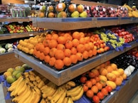 A supermarket produce section with stacked shelves displaying a variety of fresh fruits including oranges, bananas, grapes, and apples. The fruits are arranged neatly with prices labeled below each section.